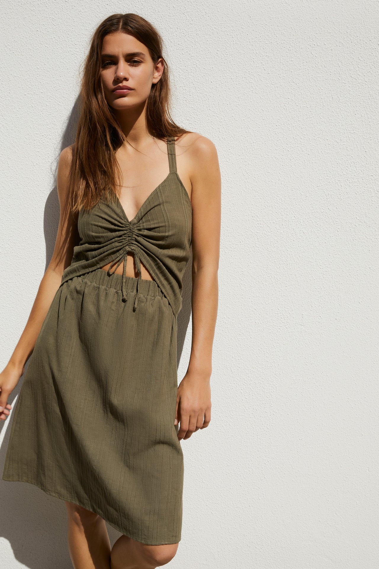 Else Ponza cotton dress in khaki green. The dress features a deep v neckline, halter style straps, a ruched front with tie closure, midriff cutout, elastic waistband, and an a-line knee length skirt. Shown on model, front view.