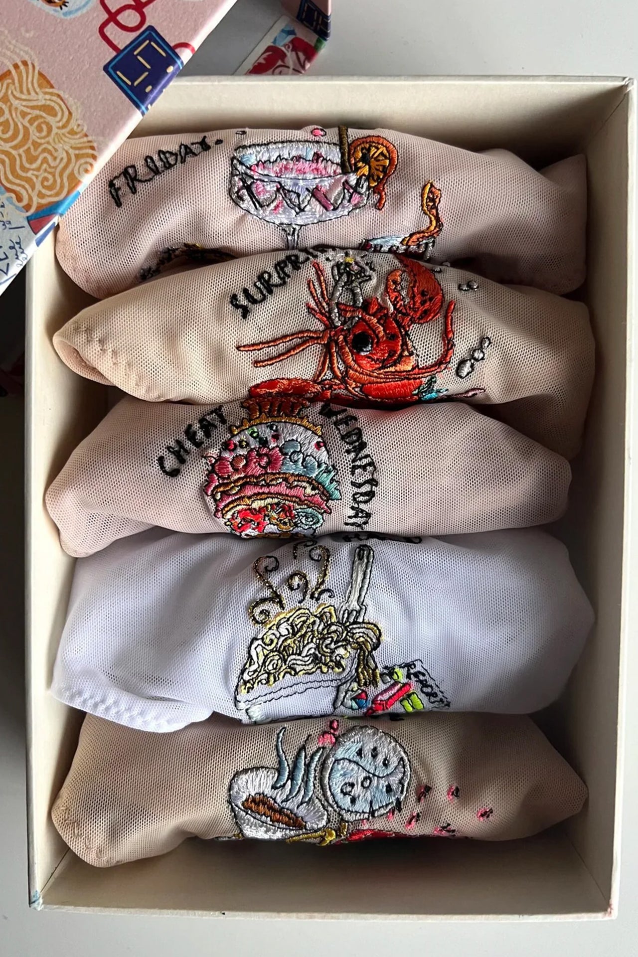 Love and Swans Days of The Week food brief box set. Five folded briefs in various colors are in a colorful gift box, seen from above. The lowest is made of beige mesh with coffee themed embroidery, the next is whte mesh with takeout embroidery, next is beige with ice cream embroidery, fourth is also beige with a lobster embroidery, "Friday" is a blush pink mesh with cocktail embroidery.