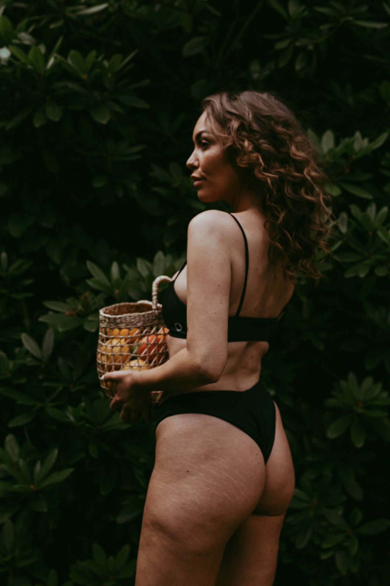 Nero black Canggu low cut swim bottom by Copenhagen Cartel. Front view on model holding a basket of mangoes and wearing matching Sanur swim top.