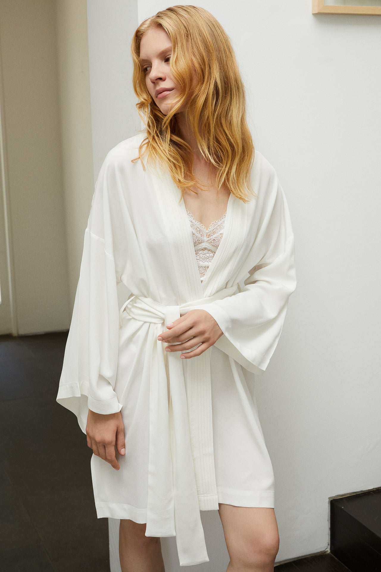 Else Diana silk robe in off white, shown on model, front view. The robe has a pleated detail along the collar, has an above the knee hemline and wide sleeves hitting just above the wrist. Shown tied at waist with a white lace bralette underneath.
