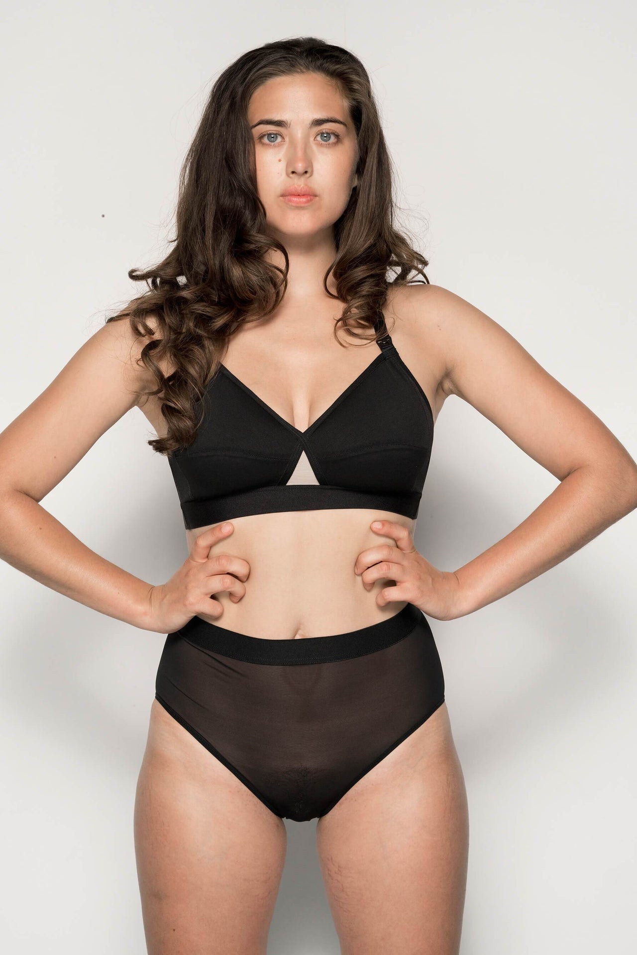 La Fille d'O Going Exact maternity/nursing bra in black with beige triangle detail at center front. Image shows v-neckline, shaping horizontal cup seams, and comfortable wide elastic under band. Plastic clips attaching cup to shoulder straps are also visible. Front view shown on model in sheer black mesh high waist briefs.