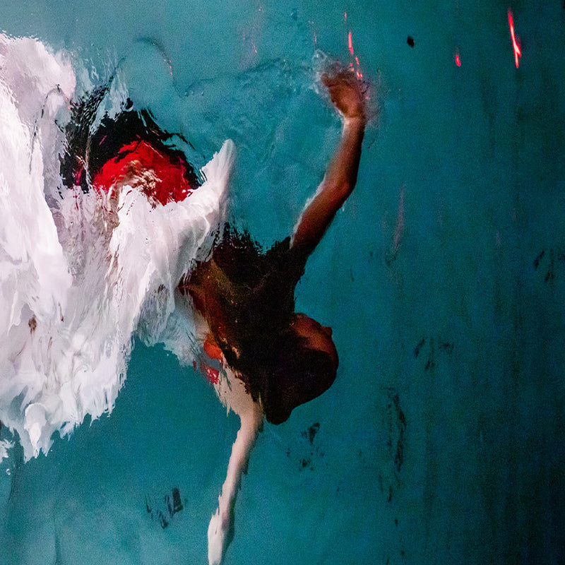 A figure swimming underwater in a bright blue pool at night, with arms outstretched and trails of white and red fabric surrounding and following her, like a water nymph or mermaid