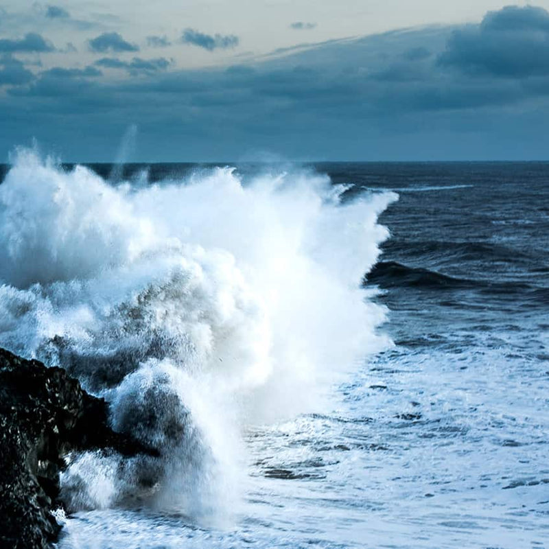 A wave crashes onto a large rock, the rest of the sea is dark and the sky looks mostly gloomy with some daylight breaking through in the distance