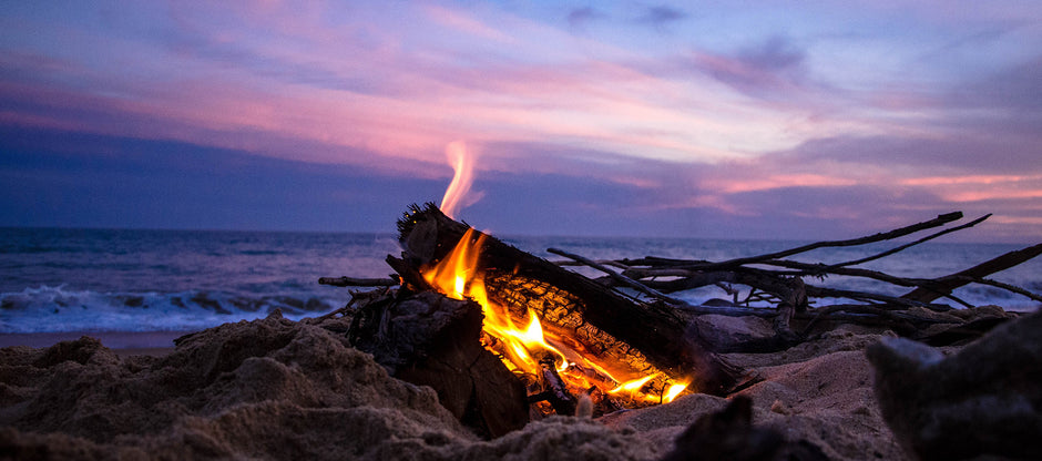 Close up of a campfire on a beach with a purple/pink/blue sunset in the background