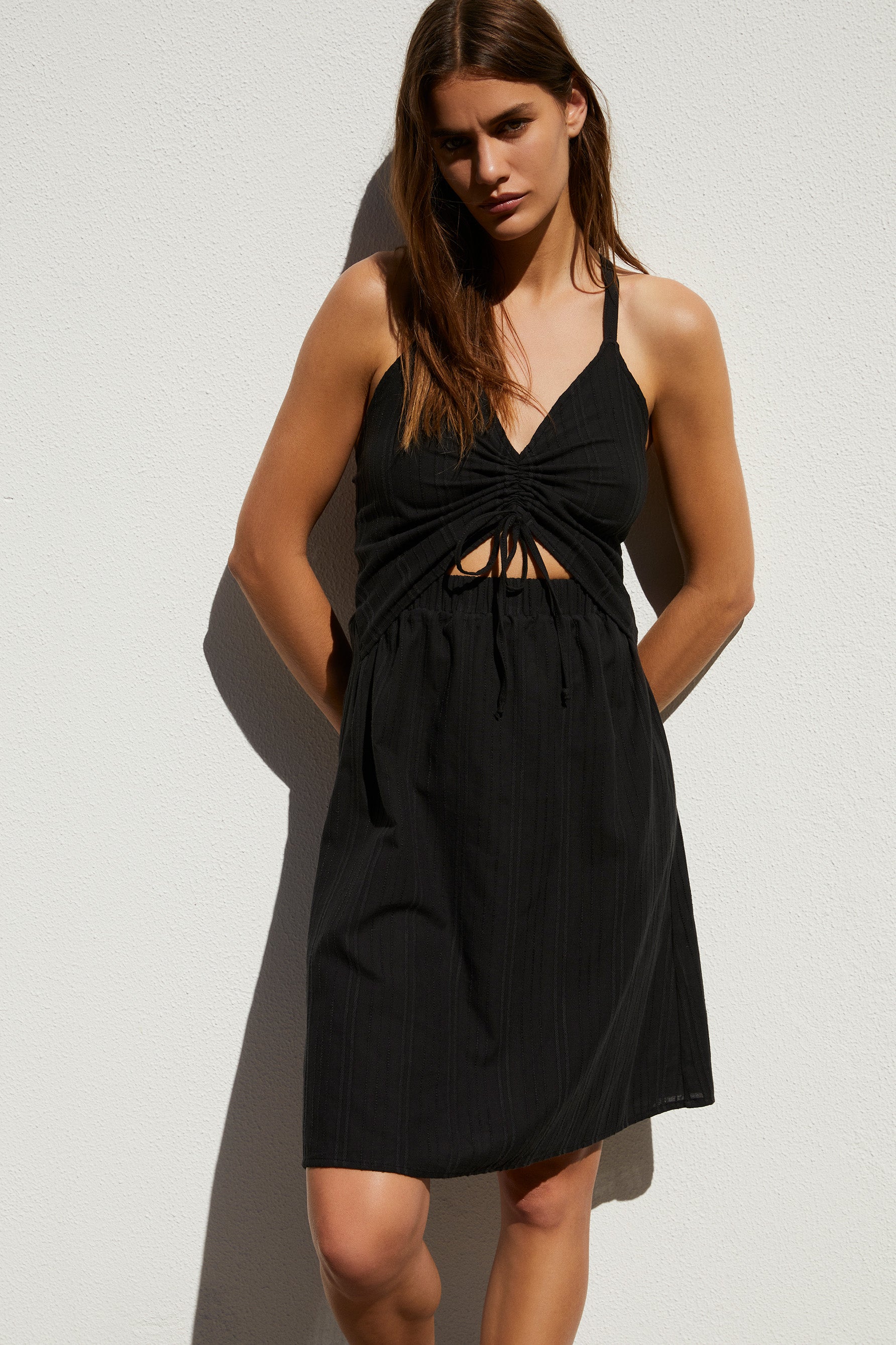 Else Ponza cotton ruched front cutout dress in black. Shown on model, front view. The dress features a v-neck ruched front with tie closure. The waistband has a cutout above the elastic. The skirt is an a-line shape that hits just above the knee. 