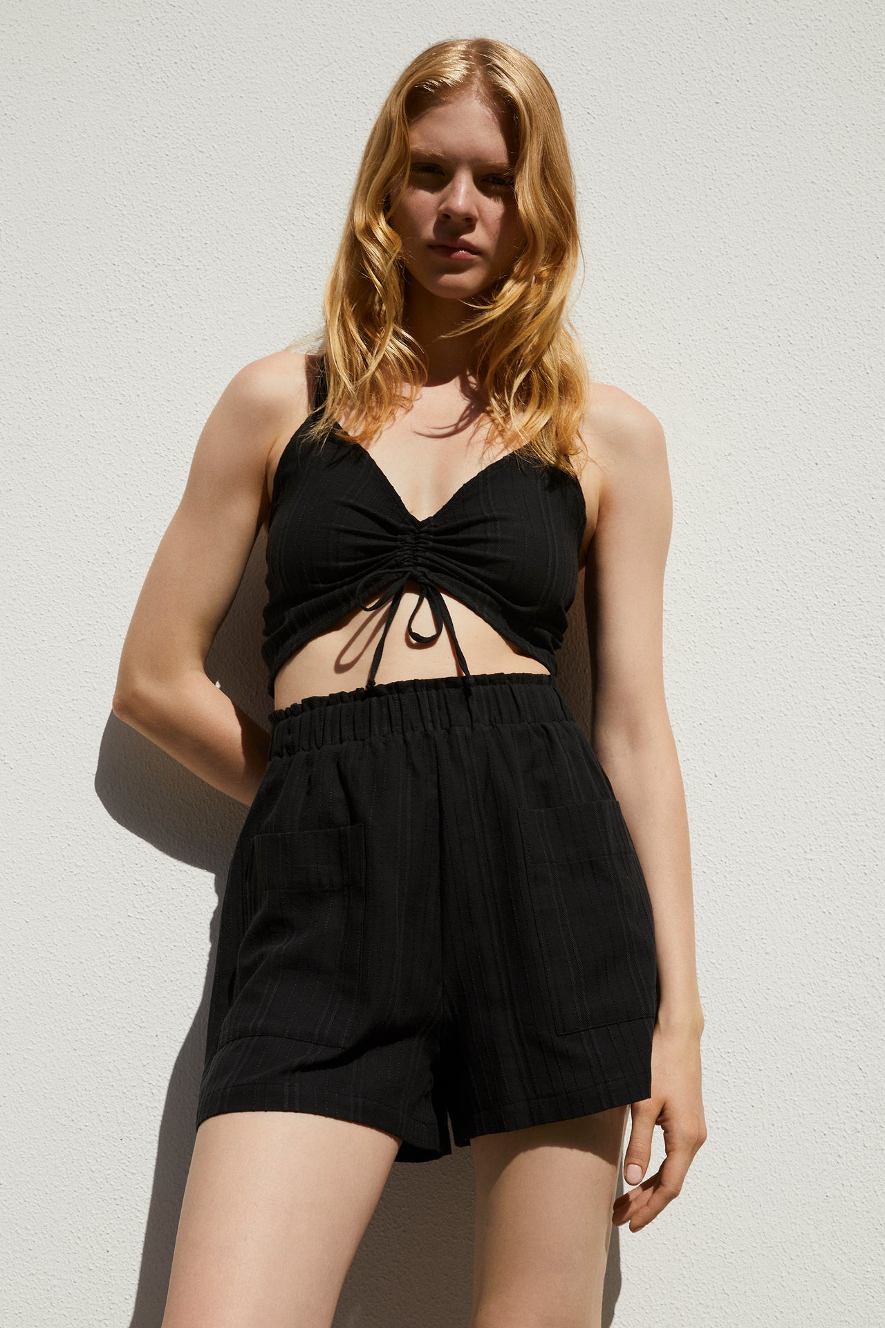 Else Ponza cotton ruched front crop top and shorts in black, back view shown on model. The top features a cross back tie closure design and the high waist mini shorts have a wide elastic waistband. Cotton fabric has a striped texture finish.