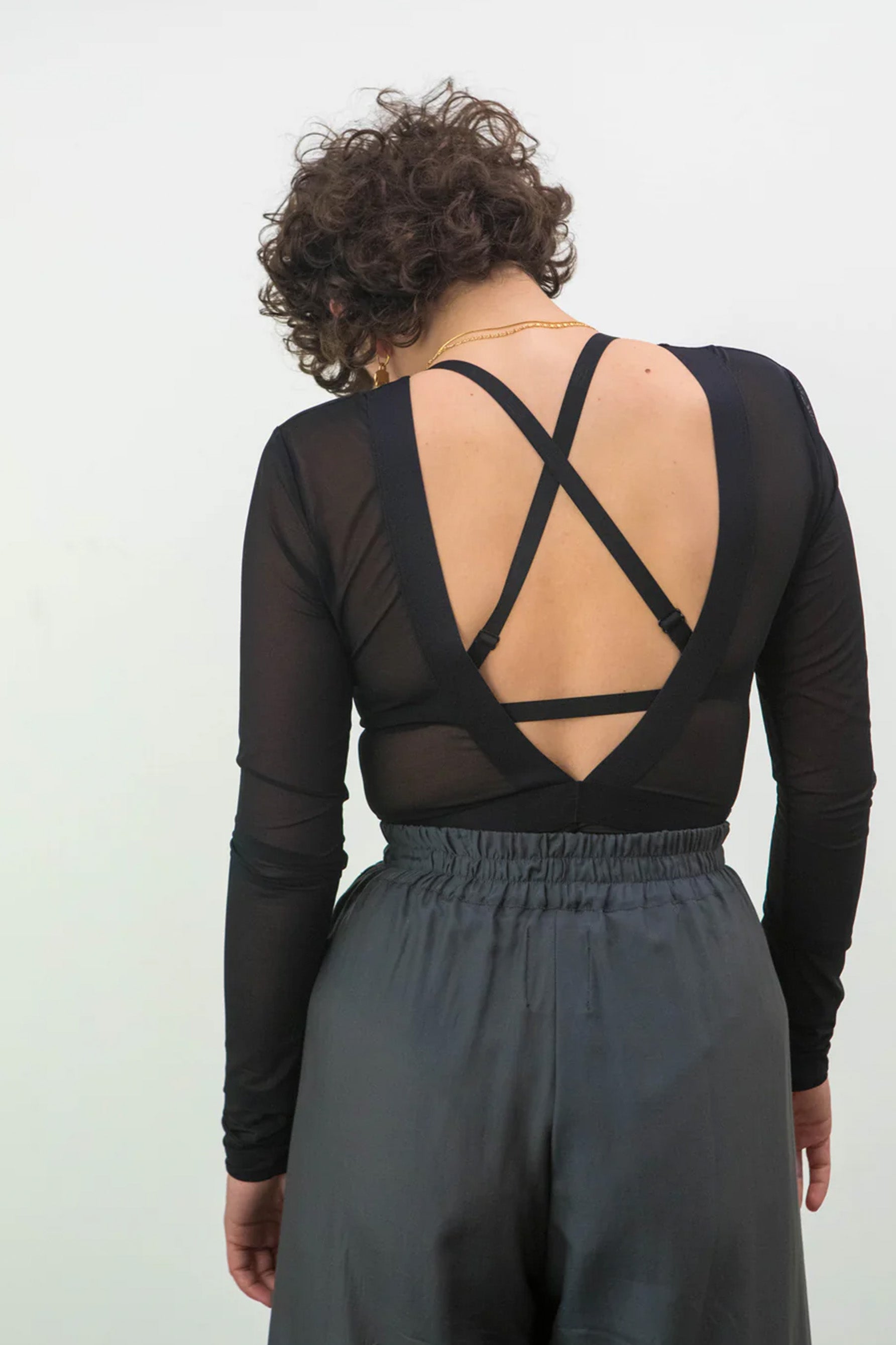 La Fille d'O Changes black swim/bra top, back view shown on model layered with deep V-back sheer mesh long sleeve bodysuit and gray blue pants. Image shows the adjustable convertible shoulder straps in the crossed position.