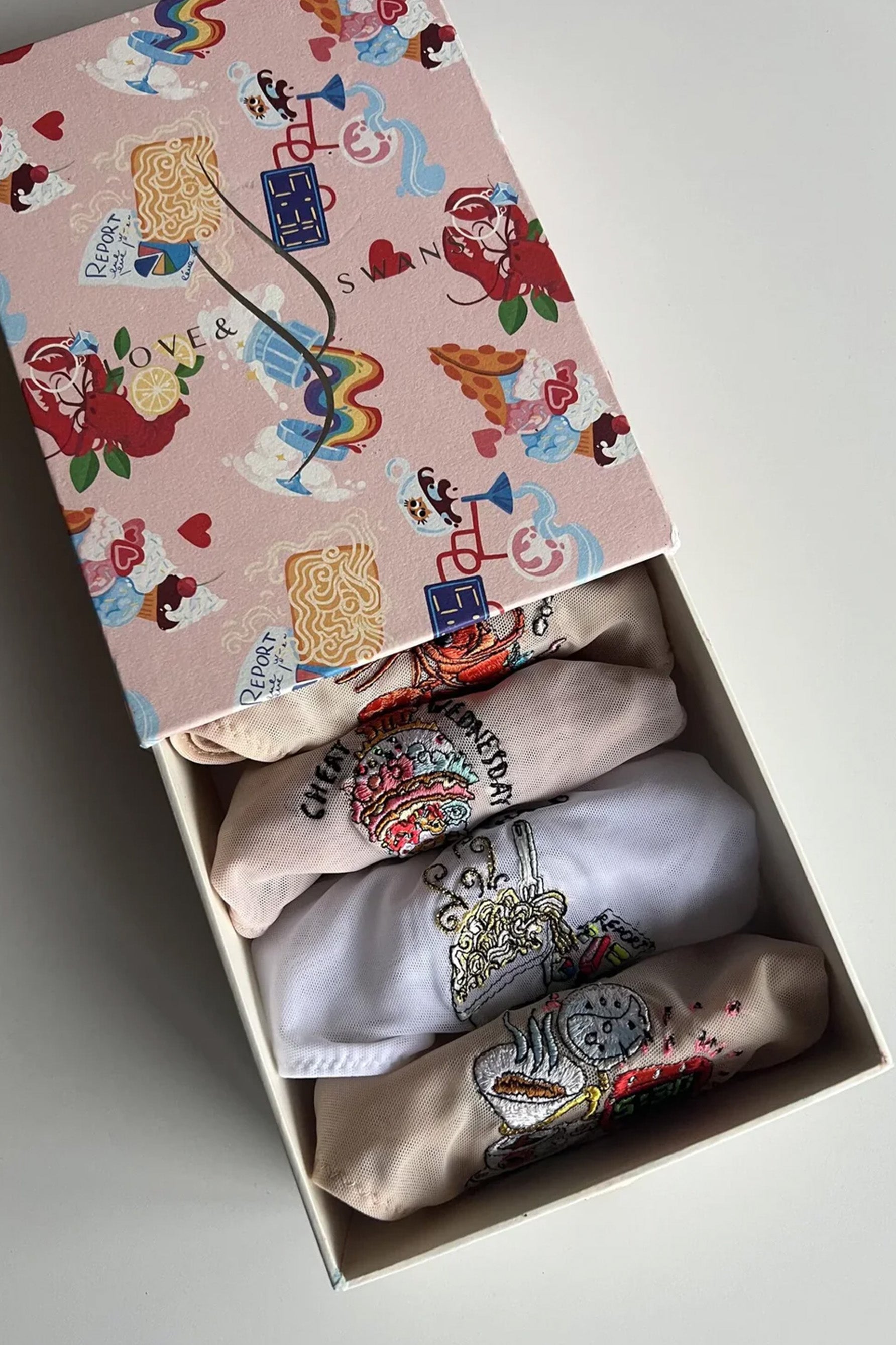 Love and Swans Days of The Week food brief box set. Four folded briefs in various colors are visible in a colorful gift box, seen from above. The lowest is made of beige mesh with coffee themed embroidery, the next is white mesh with takeout embroidery, next is beige with ice cream embroidery, fourth is also beige with a lobster embroidery.