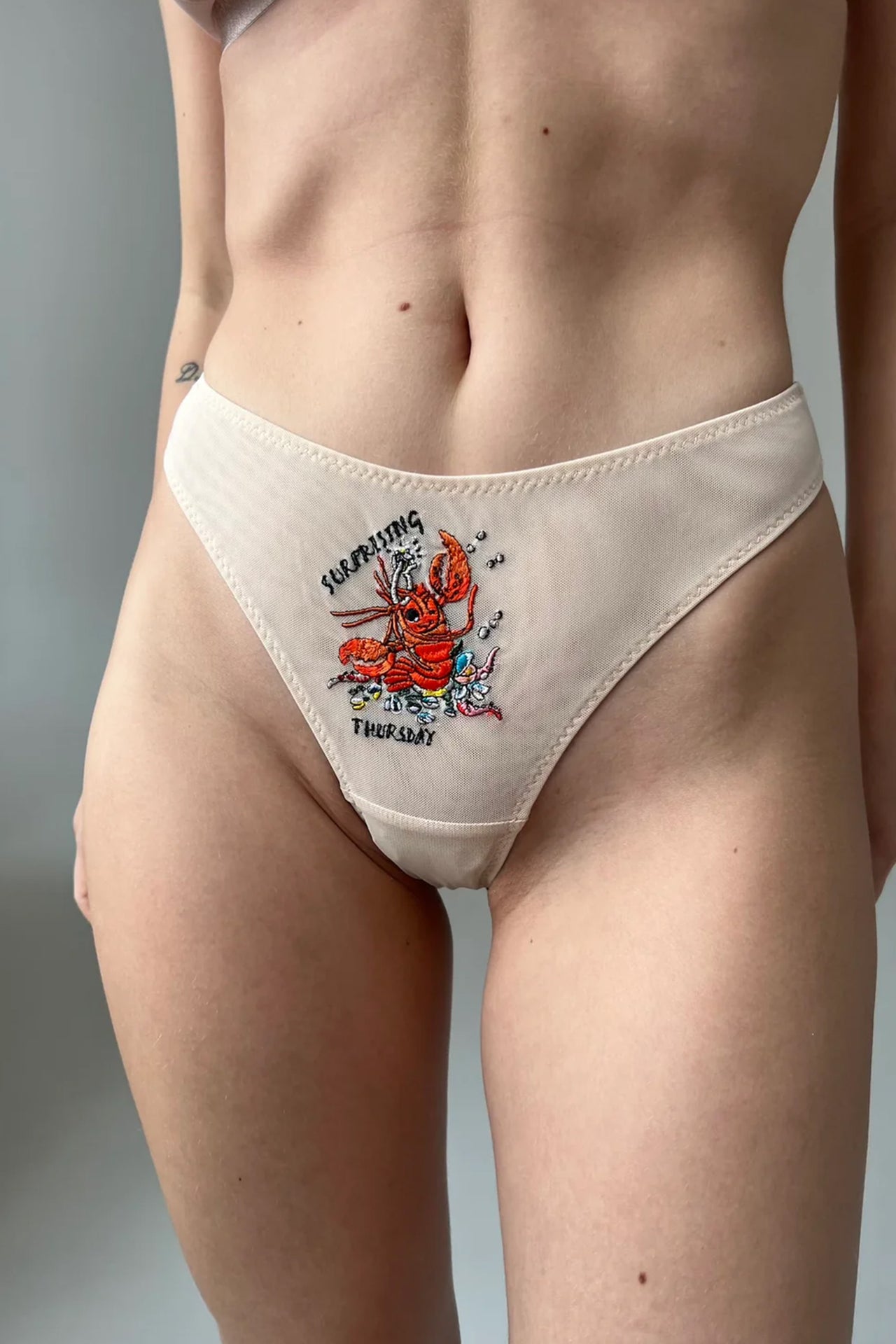 Love and Swans "Surprising Thursday" thong, front view on model. Beige mesh thong with lobster holding a diamond ring embroidered on front with the text "Surprising Thursday."