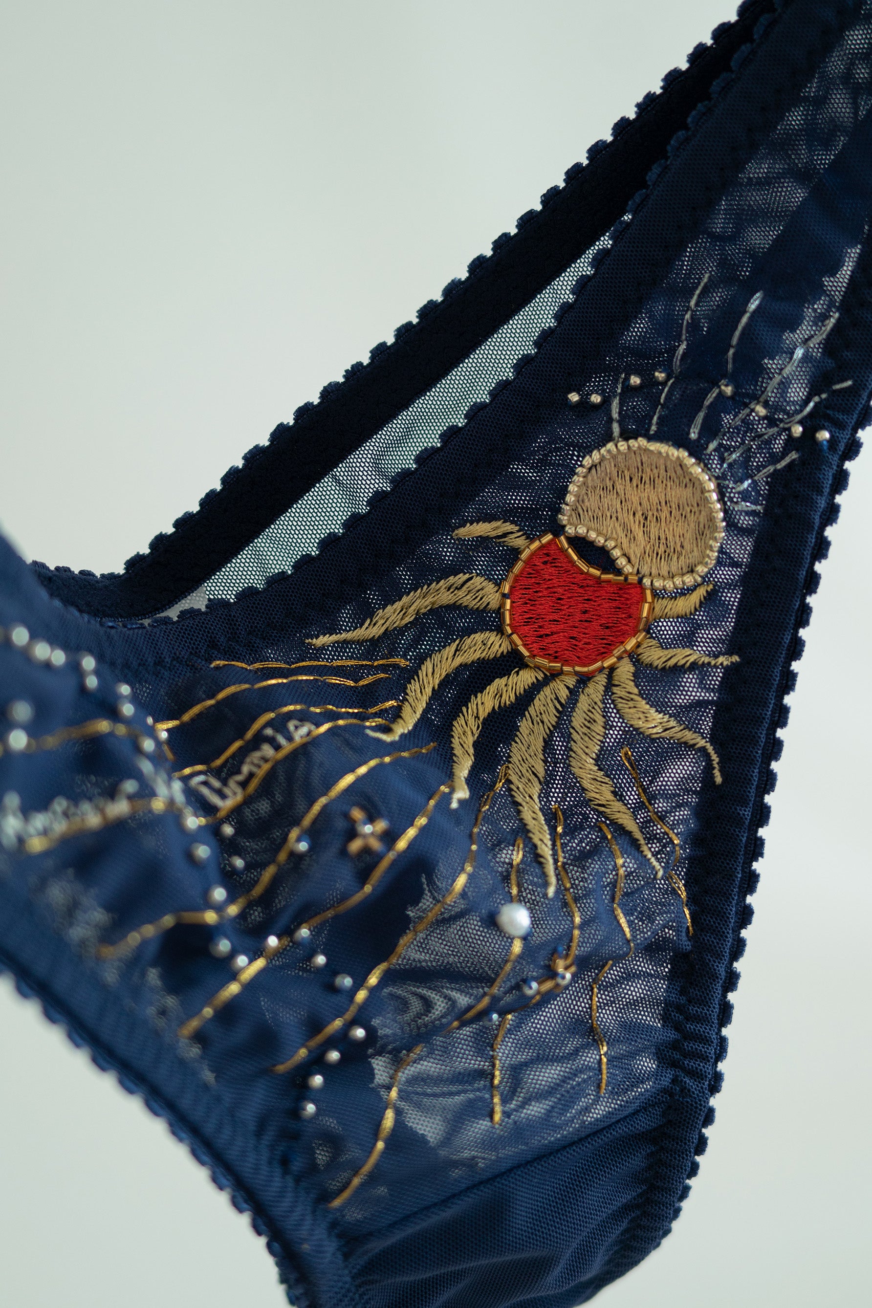 Close up view of Eclipse Thong by Love & Swans. Dark blue mesh is embroidered and beaded with a sun and moon crossing paths, rays emitting from both and the latin words for "love conquers all." 