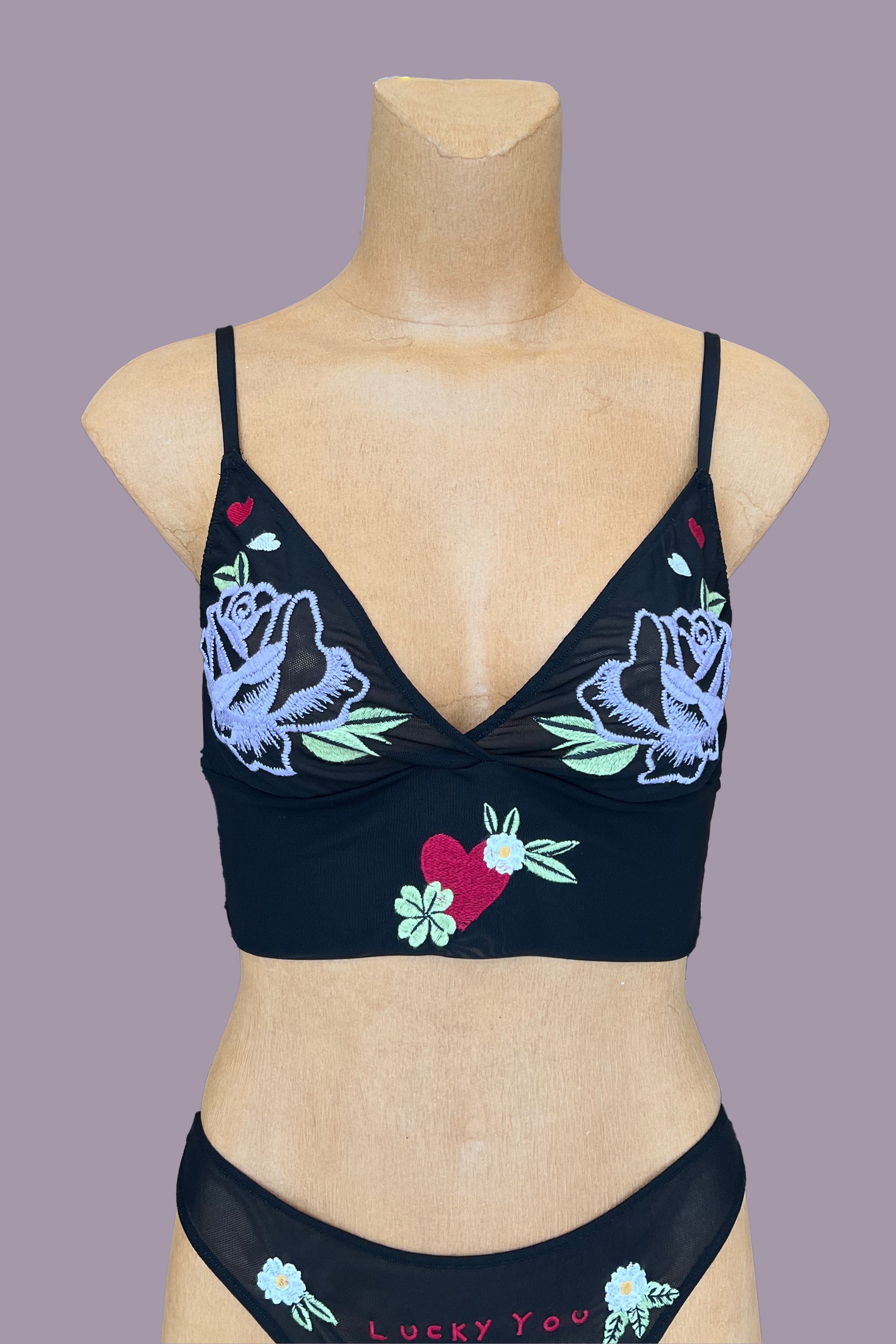 Only Hearts Aloha Honolulu Rosebud Cropped Camisole in sheer black mesh. Embroidered cups feature a purple rose with green leaves and red and white hearts. Longline band features a red heart with green and white florals surrounding it at center front. Shown on mannequin with matching "lucky you" thong. Front view.