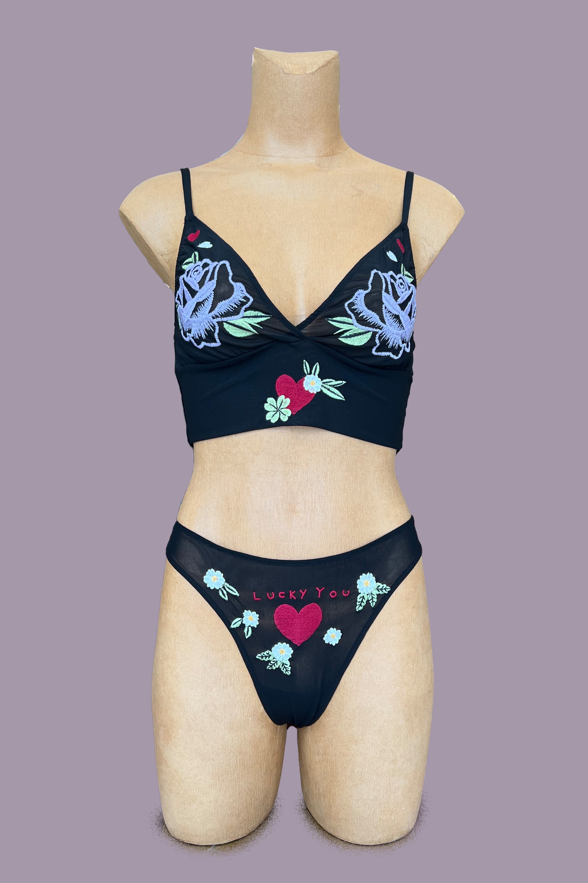 Only Hearts Aloha Honolulu Rosebud Cropped Camisole in sheer black mesh. Embroidered cups feature a purple rose with green leaves and red and white hearts. Longline band features a red heart with green and white florals surrounding it at center front. Shown on mannequin with matching "lucky you" thong. Front view.