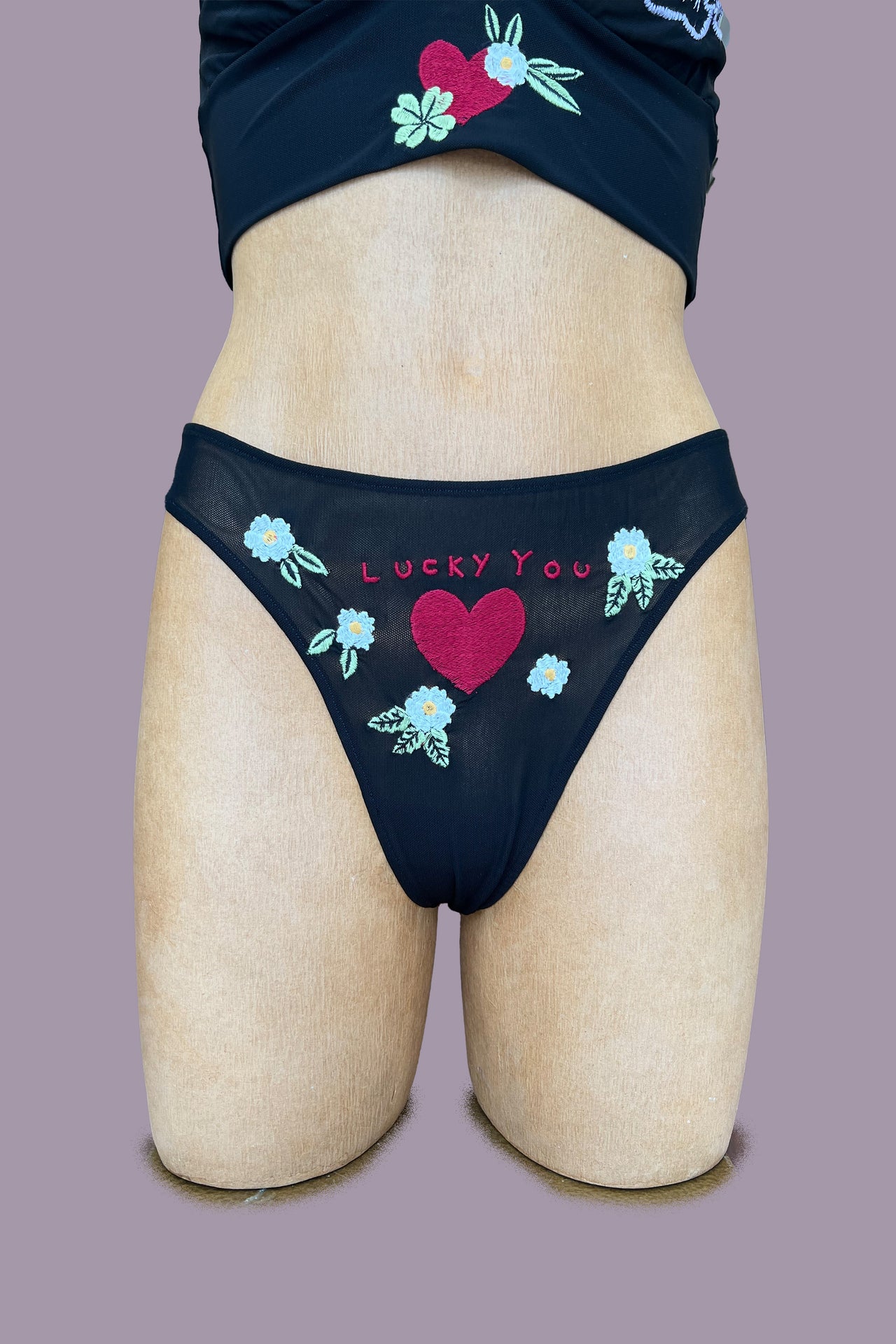 Only Hearts Aloha Vintage Thong in sheer black mesh with white and green floral embroidery surrounding and embroidered red heart and the words "lucky you." Front view on mannequin.