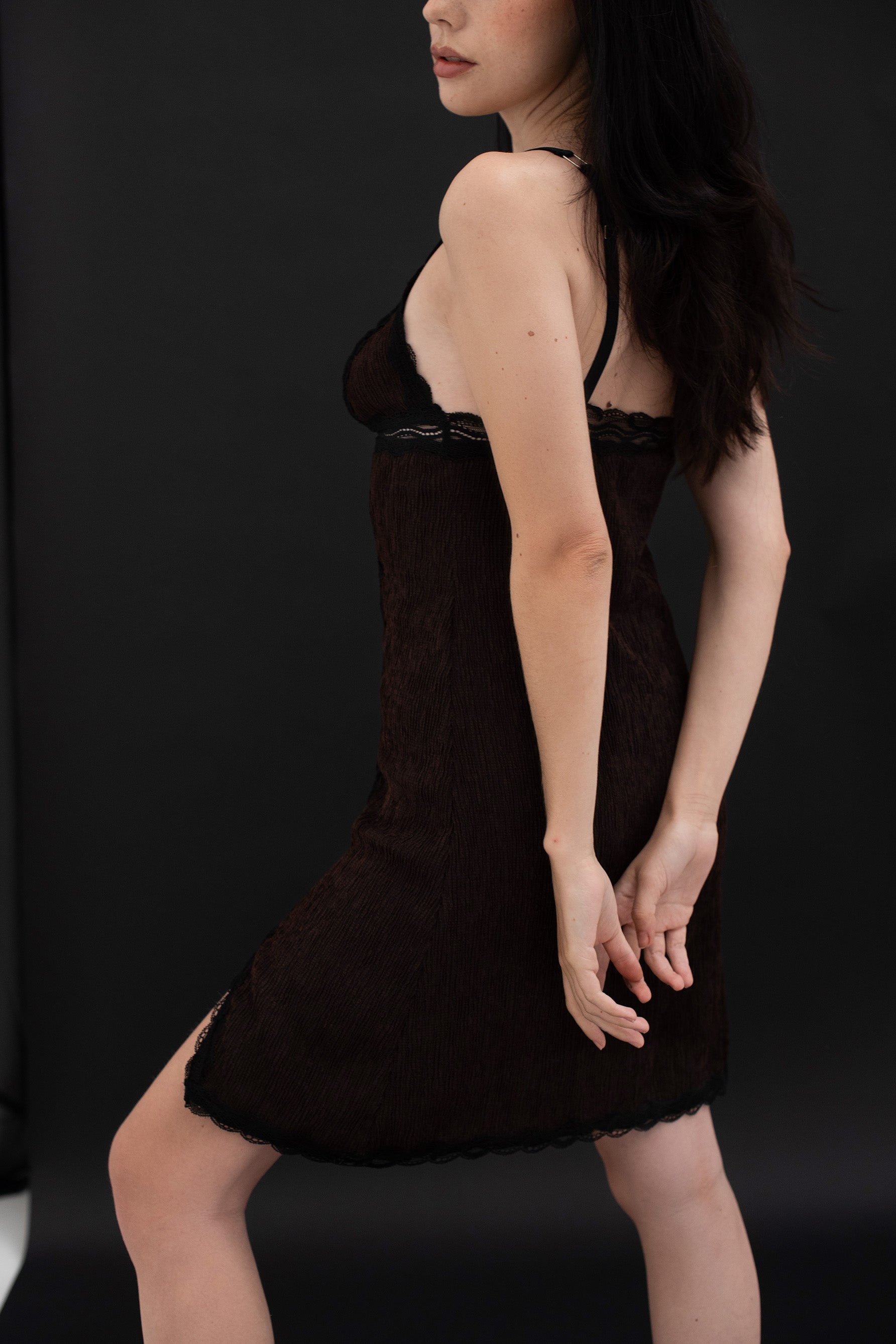 Paloma Casile La Nuit Rachel slip dress in chocolate brown and black lace. Image shows the back/side on model, including the adjustable elastic shoulder strap, lace trimmed triangle cups and sheer lace underband, along with chocolate brown pleated body fabric, and slit on front left leg. 