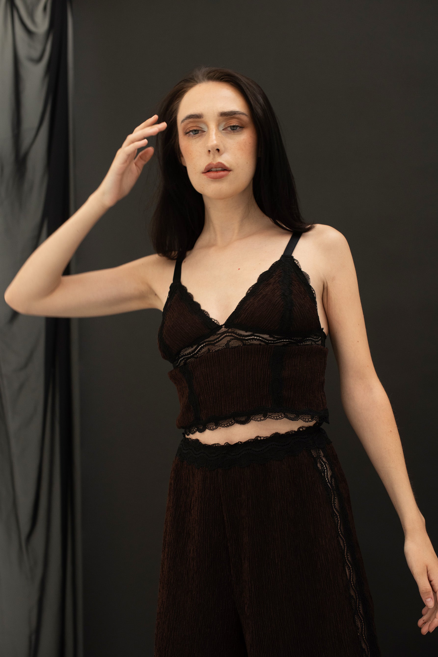 Paloma Casile La Nuit Rachel triangle cami top in dark chocolate brown and black lace, front view on model. Triangle cups with black lace trim and underband, black elastic shoulder straps and black lace trim at cropped hemline. Main fabric is brown crinkle finish. Shown with matching lounge pants. 