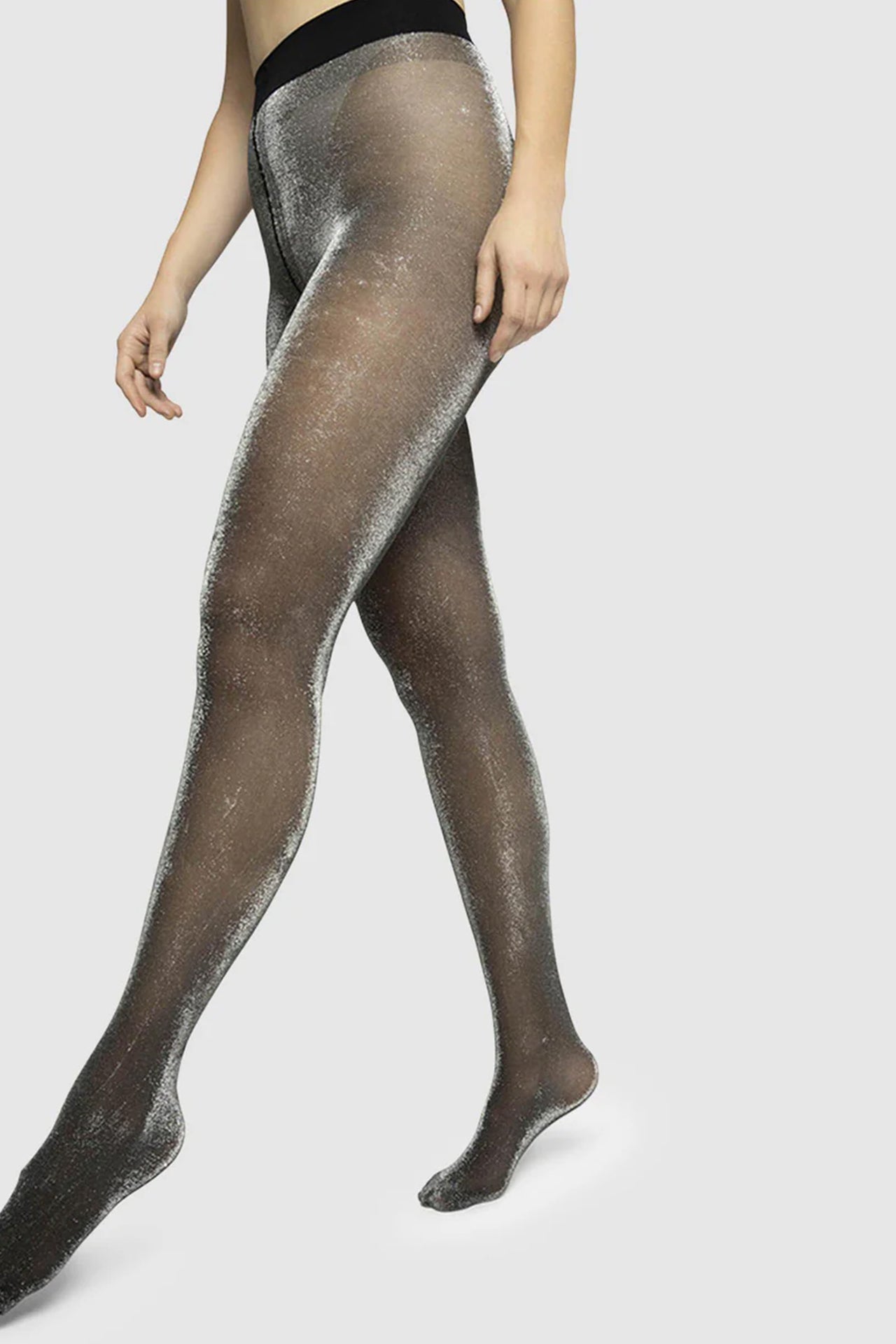 Tora Shimmery tights by sustainable hosiery company, Swedish Stockings. Detail view showing the silver glittery effect on the sheer black tights. Shown on model who is seated/squatting and resting on the balls of her feet.