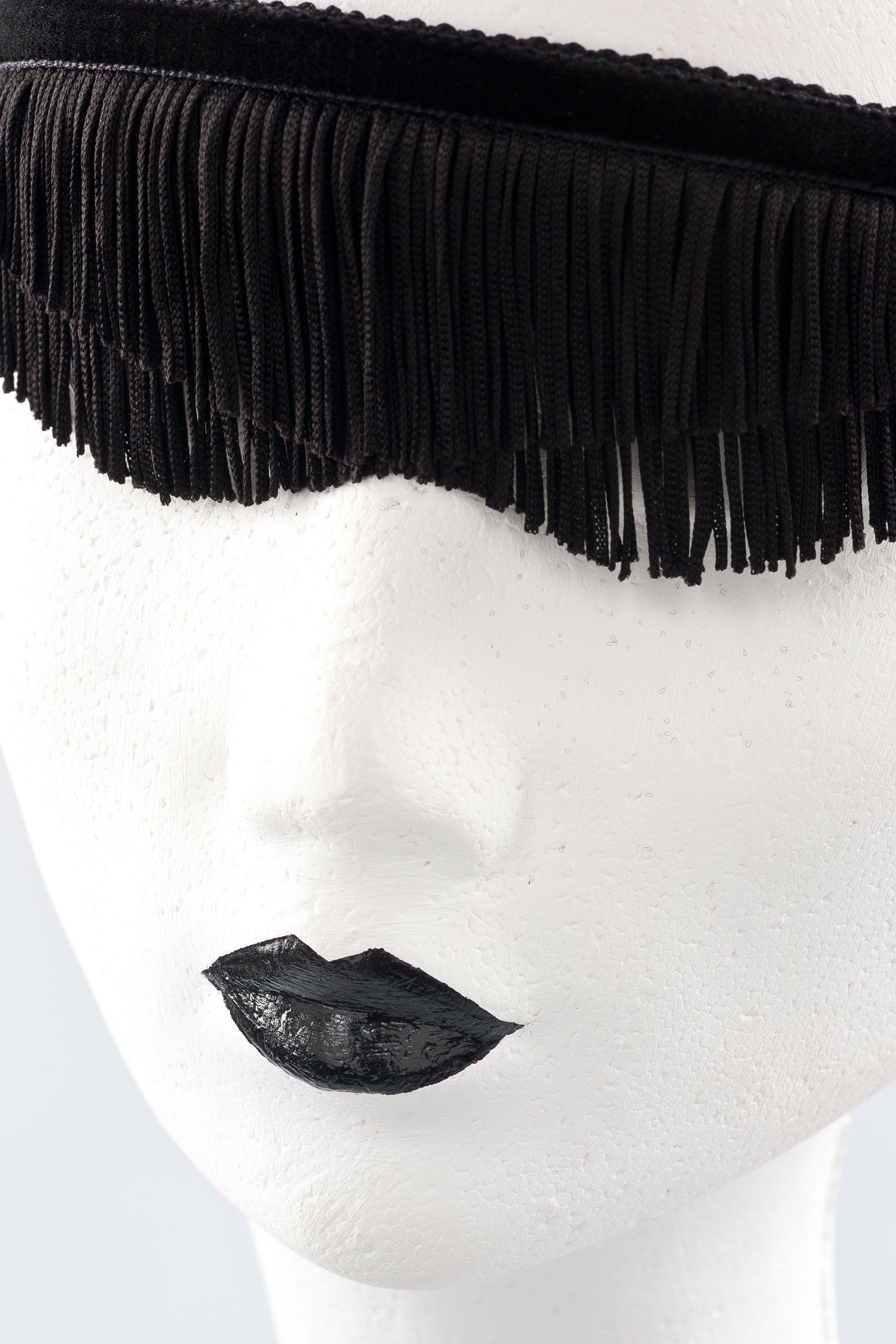 Fraulein Kink Justine fringe blindfold in black, close up front view on white mannequin. Textured fringe in two layers over the eyes bound by velvet ribbon at top. Curved finish under eyes. 