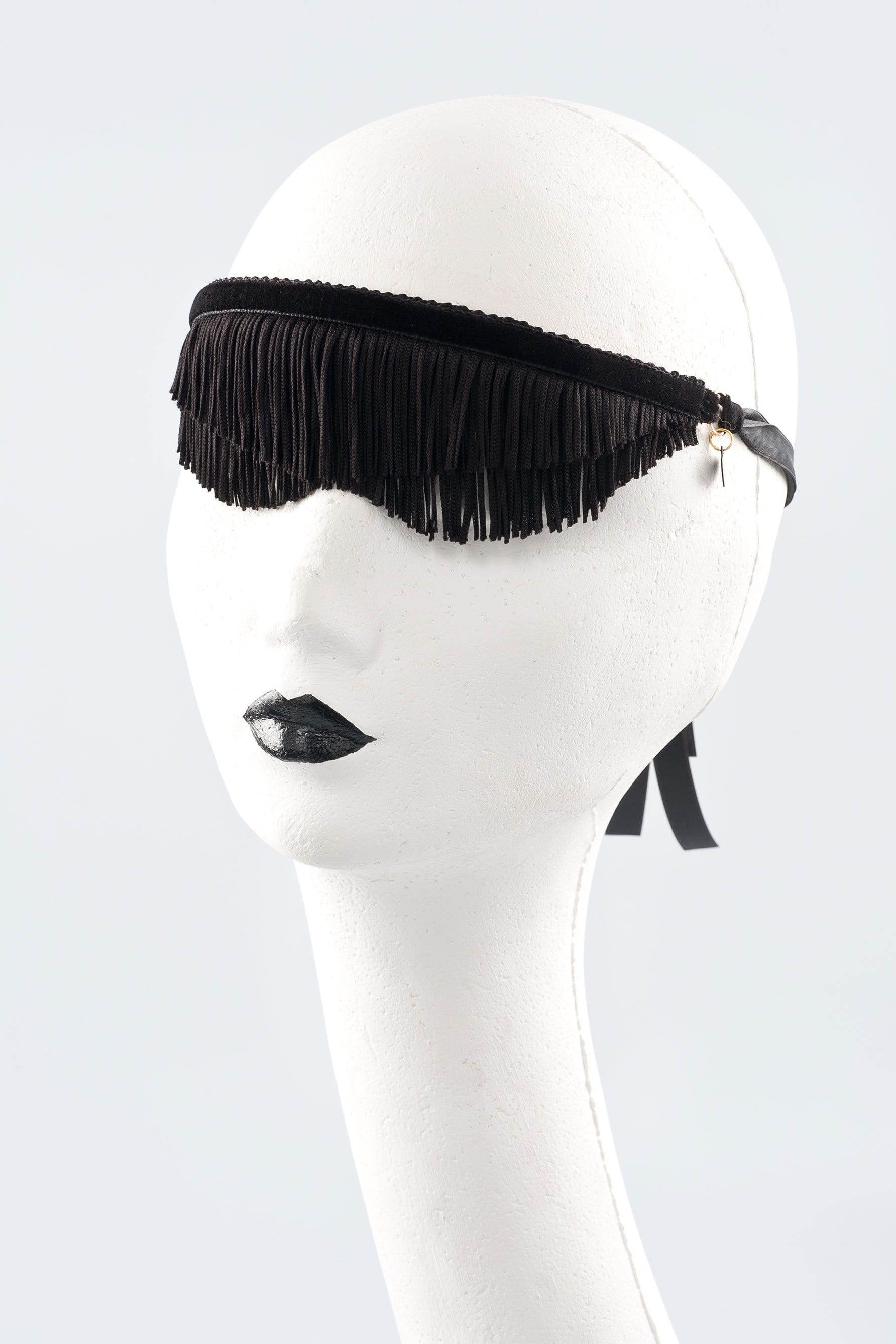 Fraulein Kink Justine black fringe blindfold, front/side view on white mannequin. Two layers of fringe bound by velvet band. Curved hem like sunglasses under eyes. Ribbon tie closure with small charm attached where ribbon meets velvet band on side of head.