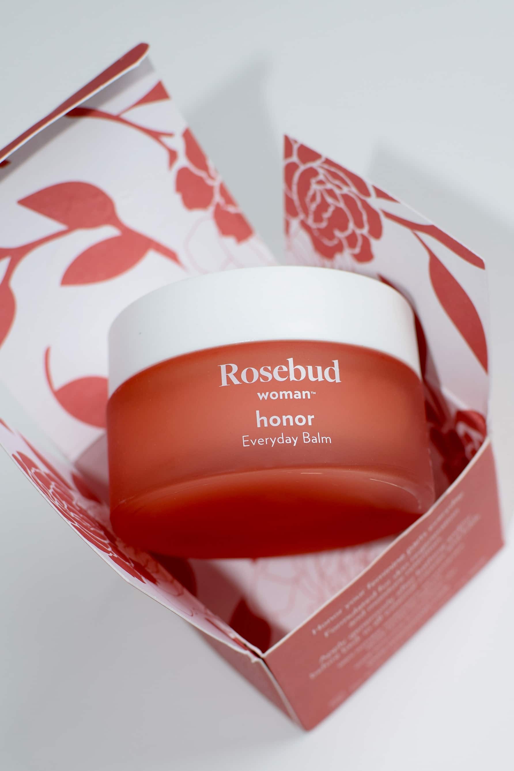 Rosebud Woman Honor Everyday Balm. Image shows the closed jar from the front, with the name of the product in white lettering on the coral pink jar with white lid. Jar is shown balancing on its open box, which is pink with white lettering on the outside, and white with a pink graphic rose print on the inside.