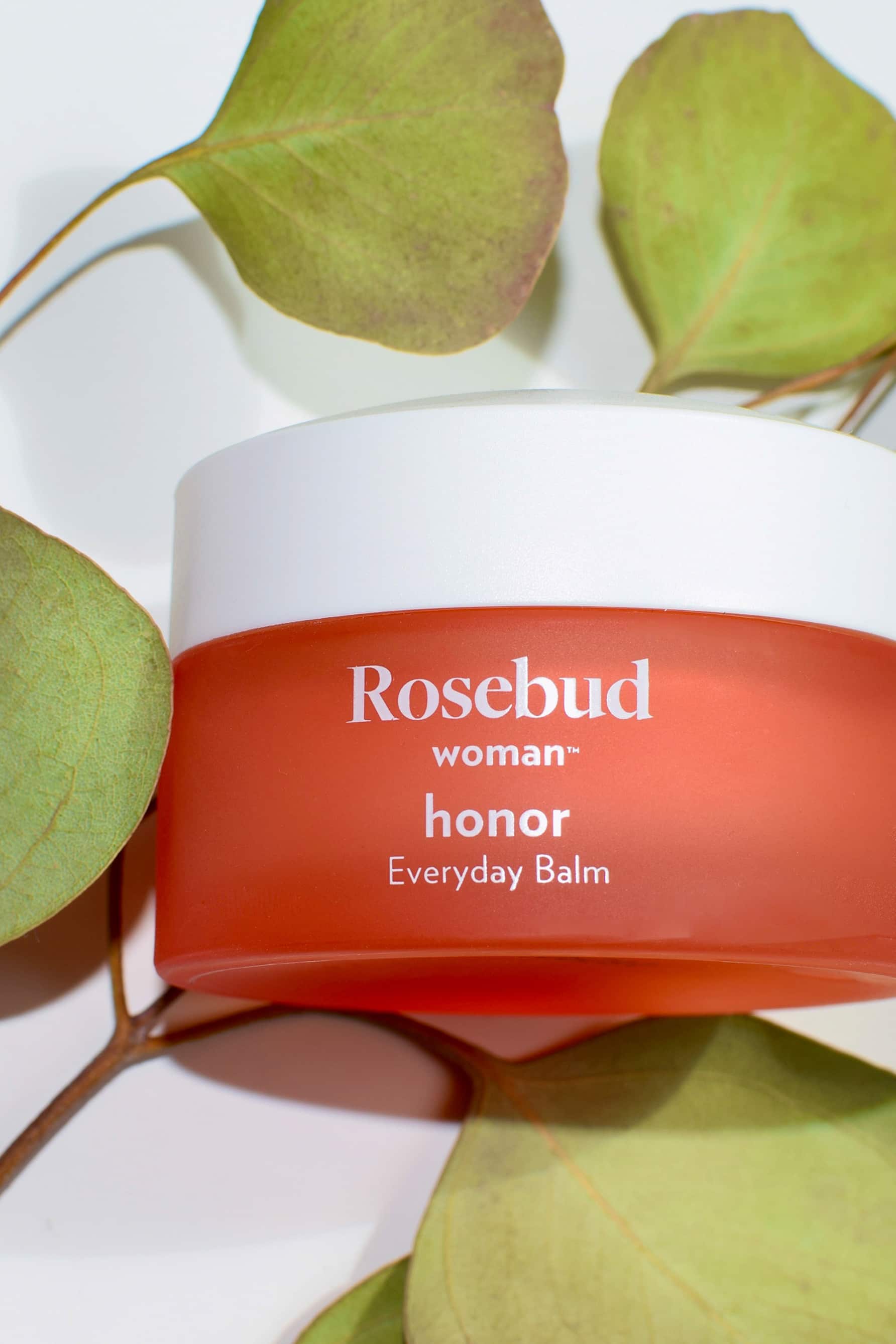 Rosebud Woman Honor Everyday Balm. Image shows the closed jar from the front, with the name of the product in white lettering on the coral pink jar with white lid. Jar is surrounded by green leaves.