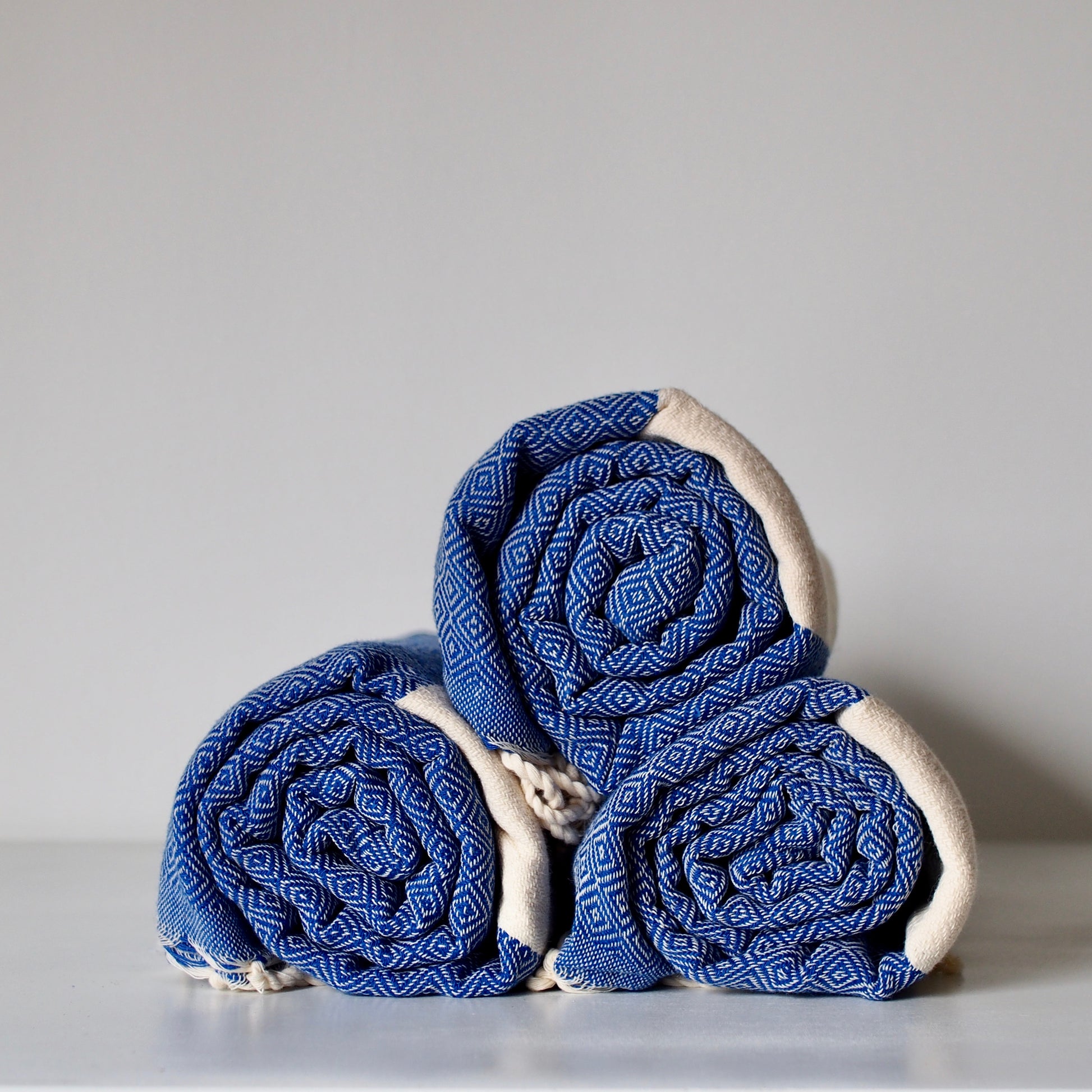 Cotton hammam towel in beige and blue diamond stripe pattern. Three towels shown rolled up and stacked in a pyramid formation. Twisted fringe ends are partially visible. 