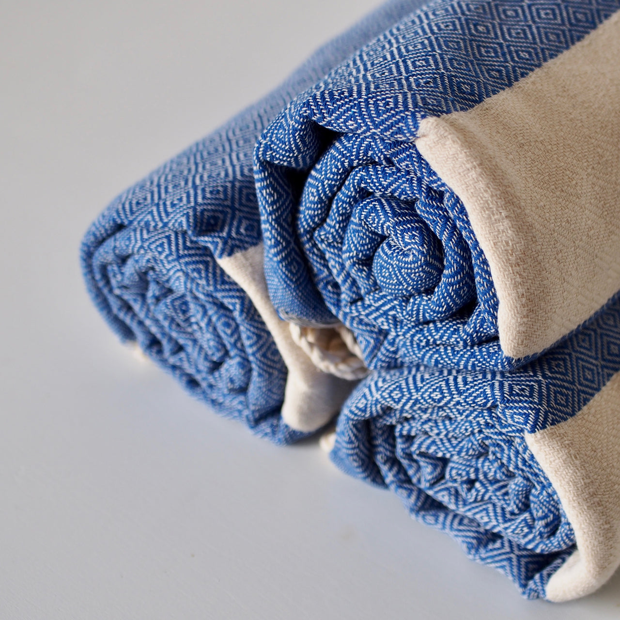 Cotton hammam towel in beige and blue diamond stripe pattern. Three towels shown rolled up and stacked in a pyramid formation. 