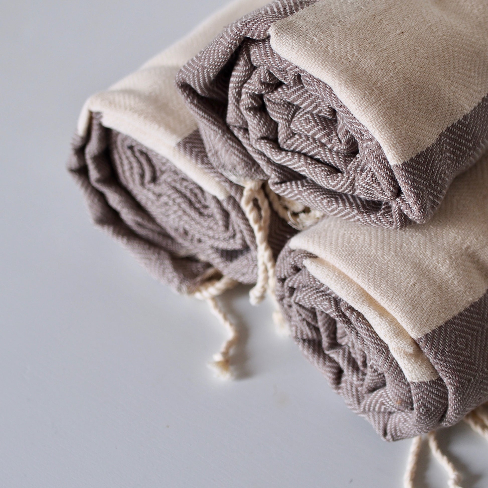 Three rolled cotton towels stacked in a pyramid shape. image shows the beige and taupe diamond shaped weaving, and fringed ends of the towels.