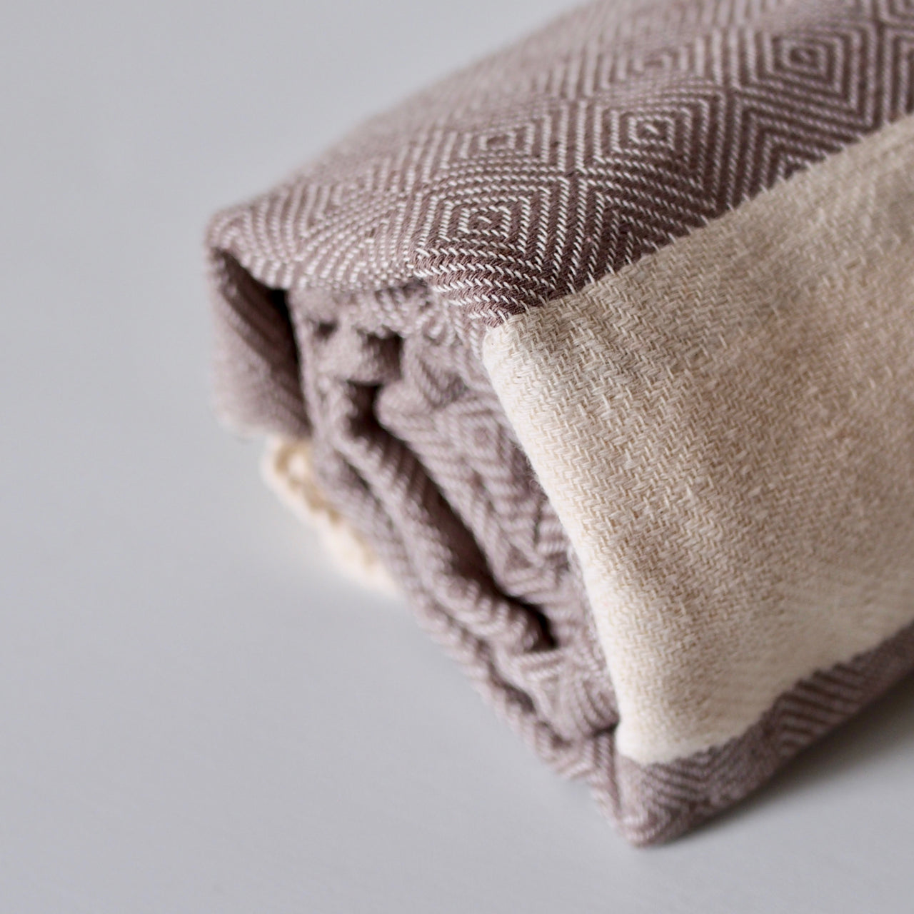 Three rolled cotton towels stacked in a pyramid shape. image shows the beige and taupe diamond shaped weaving, and fringed ends of the towels.