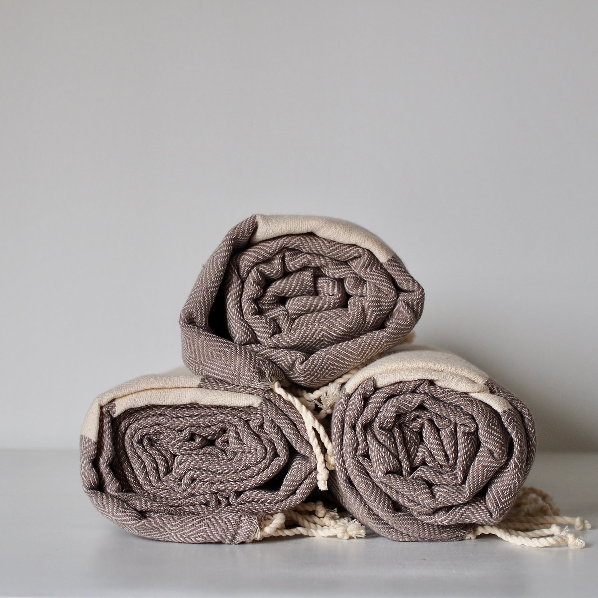 Three rolled cotton towels stacked in a pyramid shape. image shows the beige and taupe diamond shaped weaving, and fringed ends of the towels.