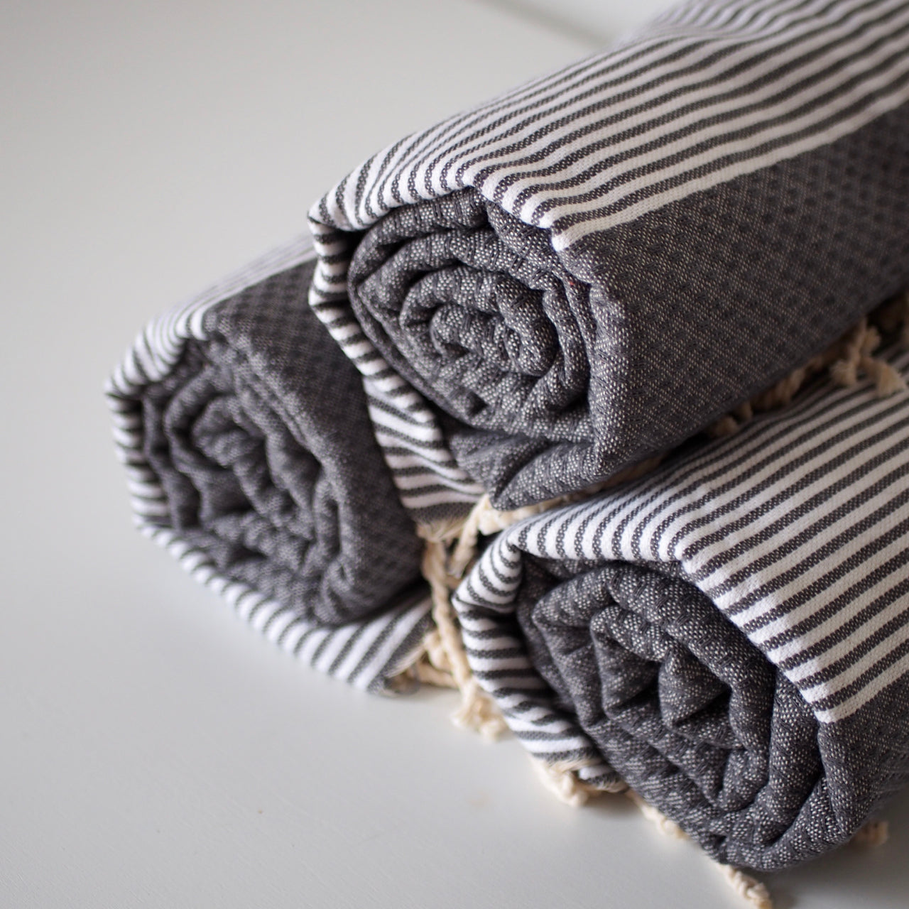 Three gray cotton hammam towels with fringed ends, rolled up and stacked in a pyramid arrangement. They are a dark gray with gray and white striped section and a waffle texture section.