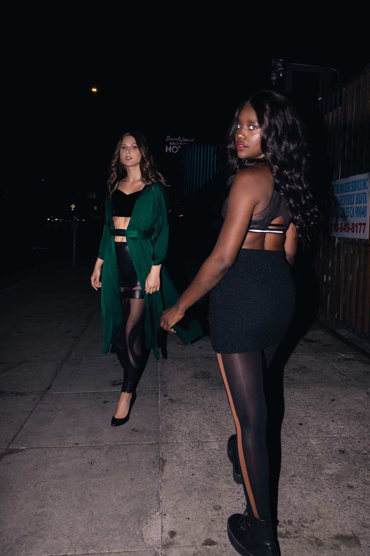 Two models standing next to each other at night on an urban sidewalk next to a fence. Right model wearing tight mini skirt and black boots with black Swedish stockings with brown stripe up the side.